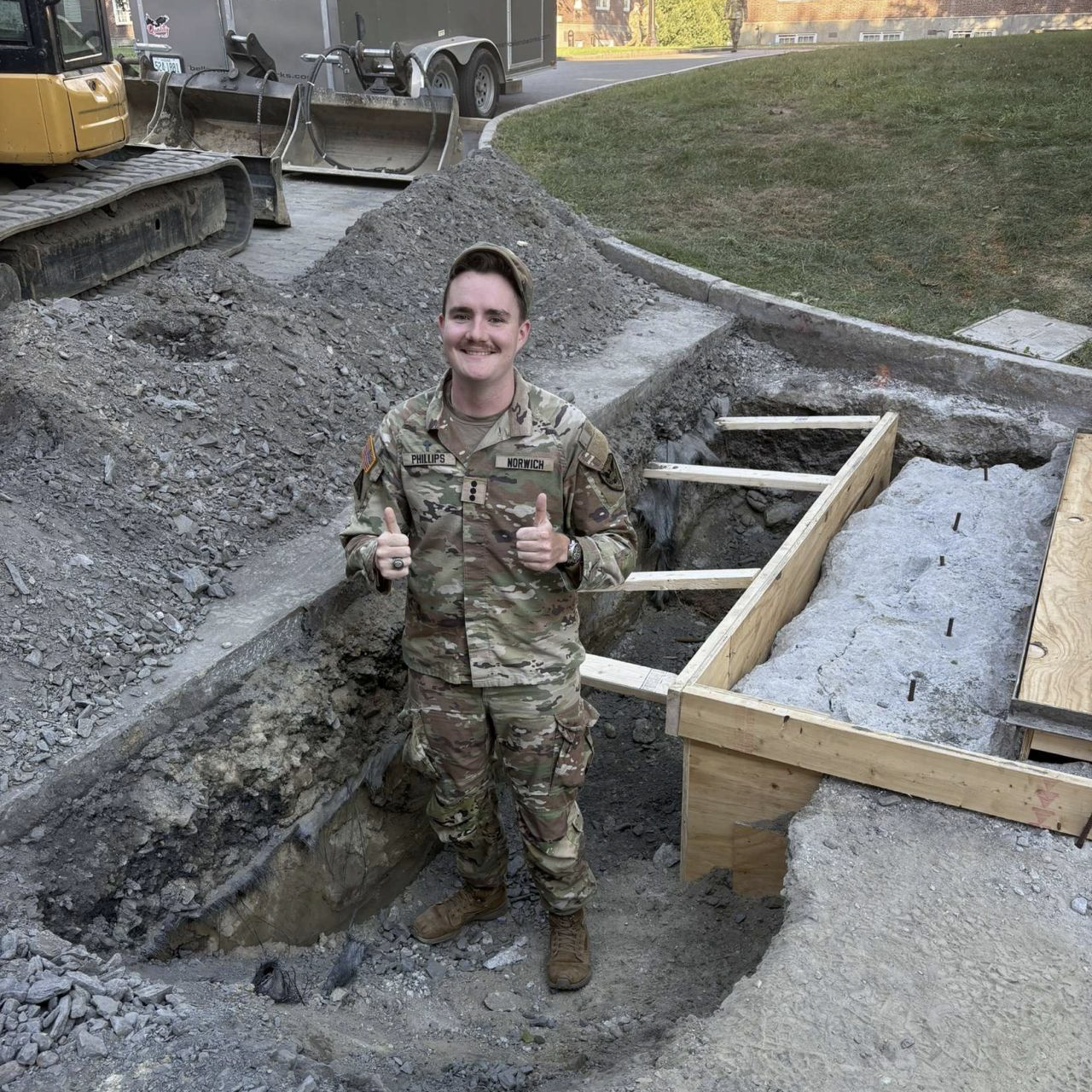 A person in military uniform stands in a shallow construction trench giving two thumbs up, with gravel piles, wooden forms, and excavation equipment nearby.