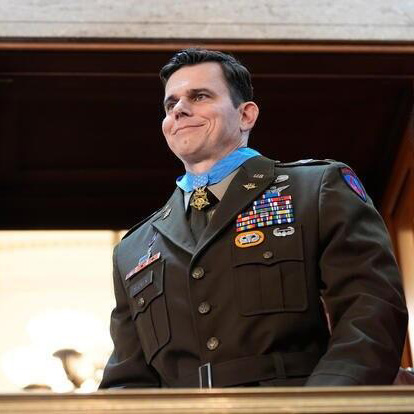 A person in a decorated military uniform stands on a balcony inside a large building.