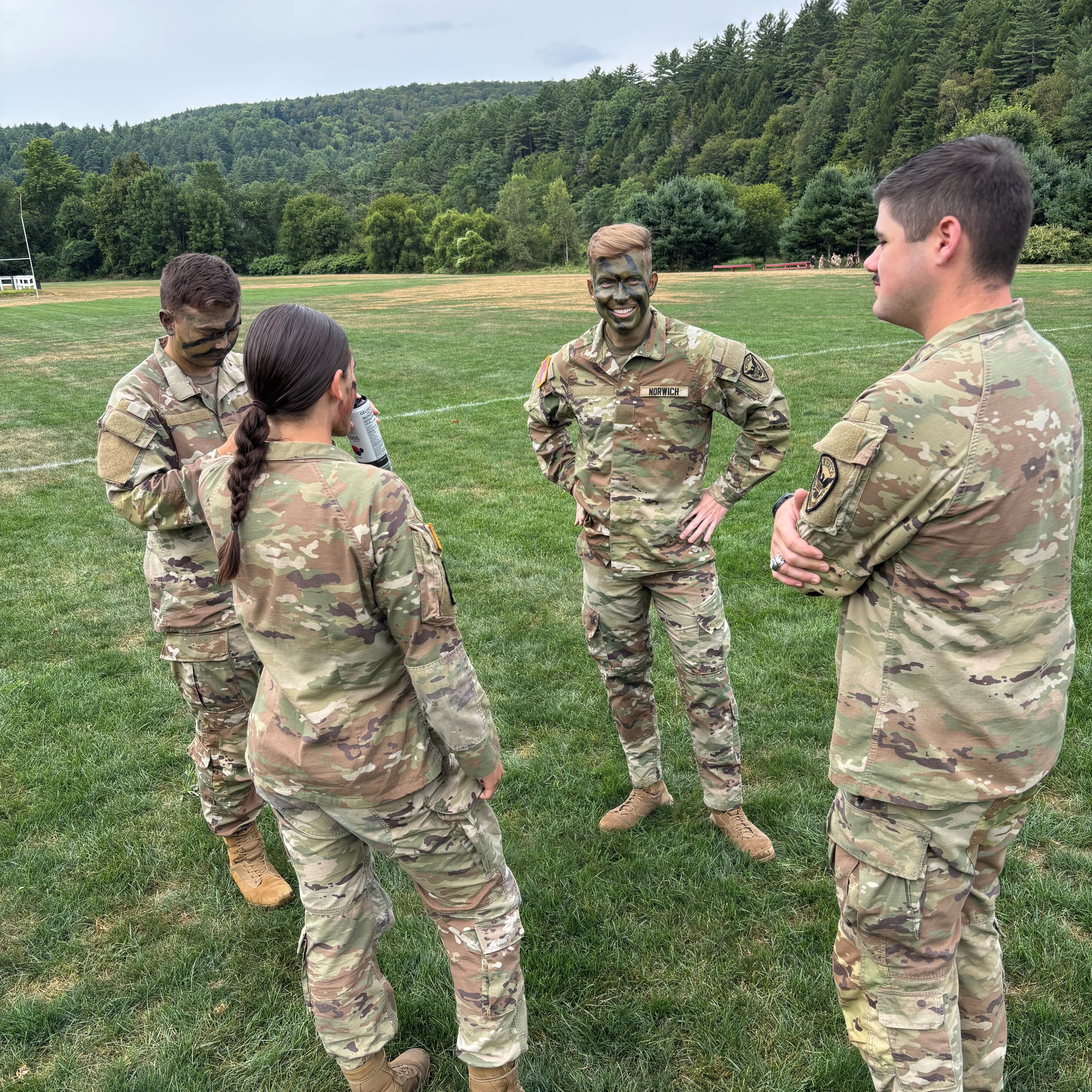 Four people in camouflage uniforms stand in a circle talking on a grassy field with trees in the background.