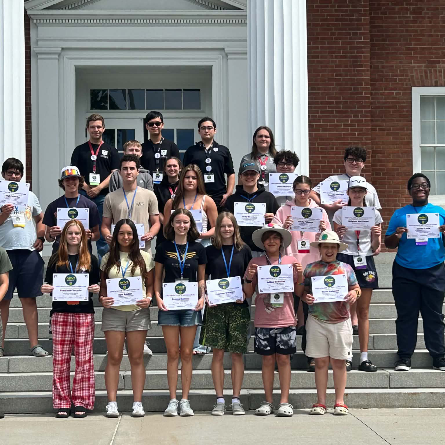 Group of students stand on building steps holding certificates in front of a brick building with white columns.