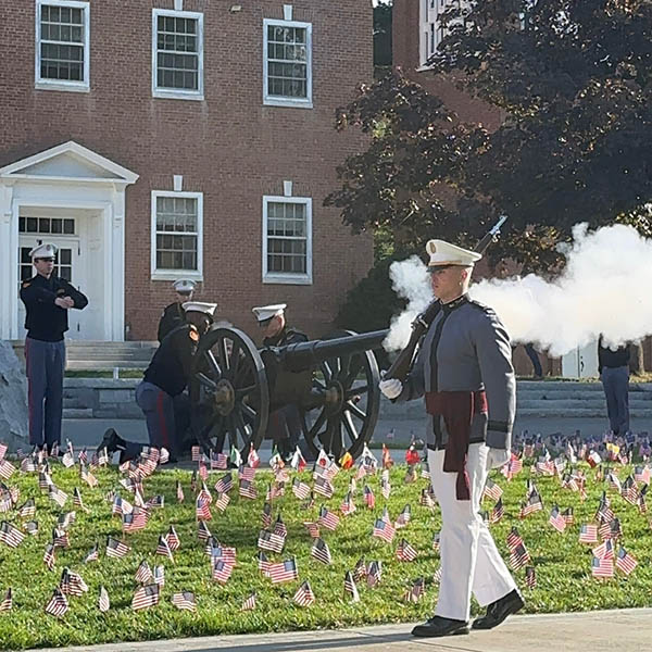 Ceremonial cannon firing by uniformed individuals in front of a brick building, surrounded by a field of small flags.