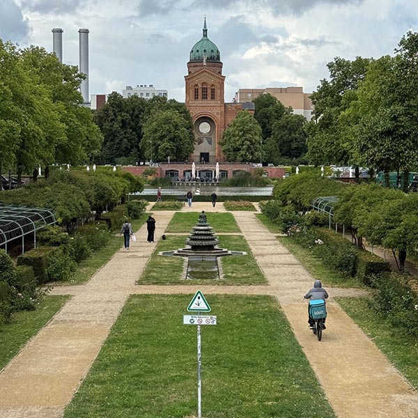 Formal garden with a central fountain, walking paths, trees, cyclists, and a brick tower in the distance.
