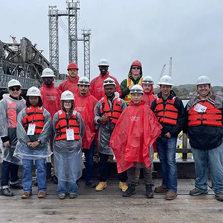 A group wearing hard hats, life jackets, and rain ponchos poses on a wooden platform beside a bridge under construction.